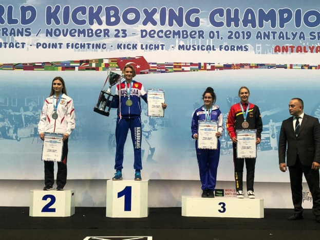 Fünf Personen halten Trophäen und Zertifikate auf einem Podium mit einem Banner "Weltweit führende Kickbox-Meisterschaft Antalya 2019" im Hintergrund.