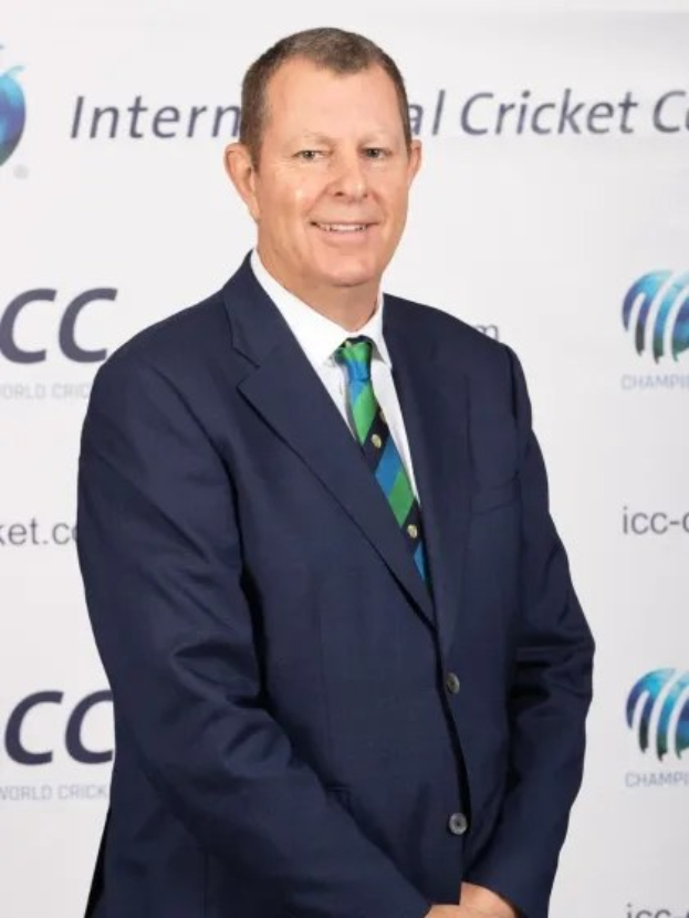 Ein Mann im Anzug und Krawatte steht lächelnd vor einer weißen Wand mit einem Banner 'ICC Cricket World Cup' dahinter.