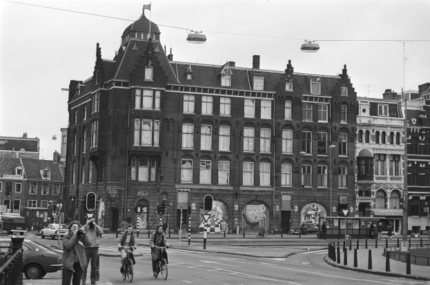 Schwarz-weißes Foto von Menschen auf Fahrrädern auf einer Stadtstraße mit Gebäuden, Straßeninfrastruktur, Kraftfahrzeugen, Fußgängern und Himmel.