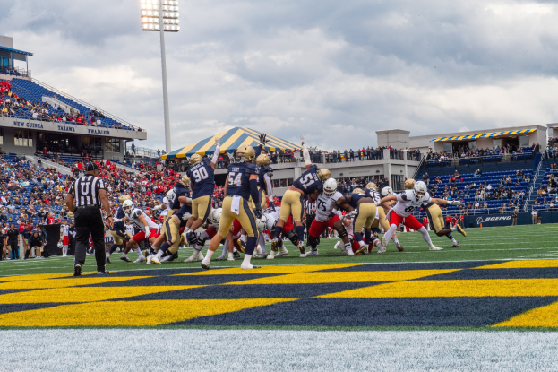 Fußballspieler in Helmen und Sportbekleidung konkurrieren auf einem Feld mit Zuschauern im Hintergrund, Stadionstrukturen und Text unten.
