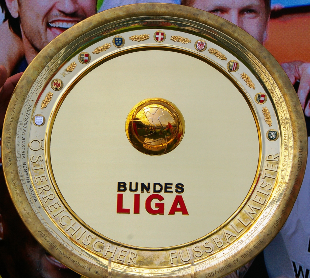 Ein Mann, der einen goldenen Schild mit der Aufschrift 'Bundesliga' hält, mit einem Banner im Hintergrund, das drei Personen zeigt.