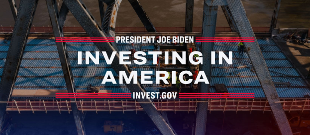 Präsident Joe Biden in der Nähe einer Brücke mit der Aufschrift "Investing in America" in der Mitte und verstreuten Gegenständen auf dem Boden.