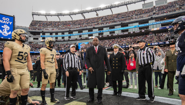 Gruppe von Männern auf einem Fussballfeld mit einer Stadionkulisse im Hintergrund, ein Bildschirm zeigt "New England Patriots" und eine Person mit einer Kamera unter Helmen.