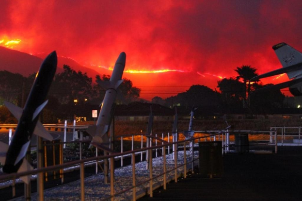 Ein Flugzeug fliegt über eine Start- und Landebahn mit Raketen, umgeben von Zäunen, Mülltonnen, Bäumen, Laternenmasten und Gebäuden, mit Rauch von den Kalifornischen Waldbränden im Hintergrund.