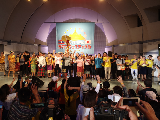 Gruppe von Menschen auf einer Bühne während einer Festival-Eröffnung, einige halten Kameras oder Telefone und andere Musikinstrumente, mit einem Banner und Deckenleuchten im Hintergrund.