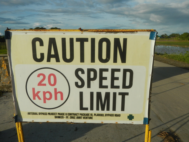 Caution speed limit sign on the side of a road with a wall, grass, water, trees, and a cloudy sky in the background.