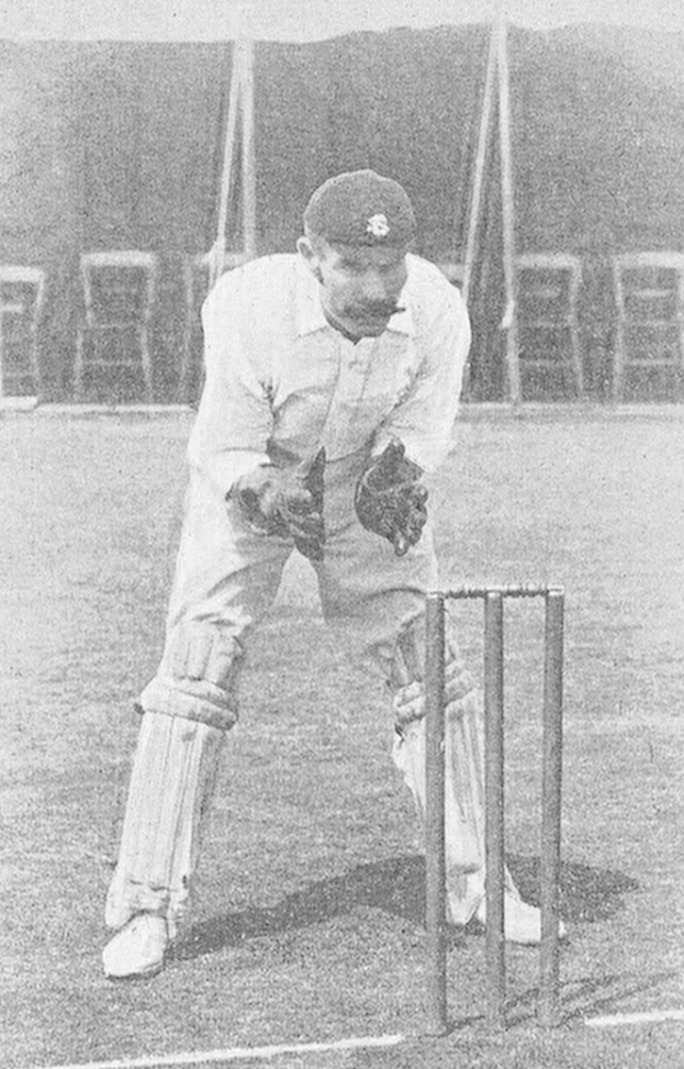Schwarzes und weißes Foto eines Mannes in Cricket-Ausrüstung auf einem Feld mit einem Wicket, der eine Mütze, Handschuhe und Schienbeinschoner trägt, mit Stühlen und Pfosten im Hintergrund.