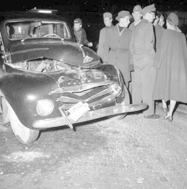 Black and white image of a severely damaged car on the roadside with a group of people inspecting it.