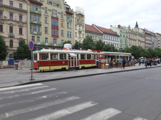Rote und gelbe Tram auf einer Stadtstraße in Prag mit hohen Gebäuden, Bäumen, einer Gruppe von Menschen auf dem Gehweg und einem Wolkenhimmel sowie einem Schild an einem Pfahl.