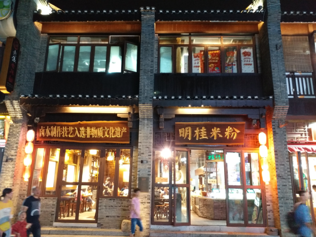 Eine Gruppe von Menschen geht an einem hell erleuchteten Gebäude mit Glasfenstern und -türen vorbei, in dem sich augenscheinlich ein Restaurant mit Gästen befindet.