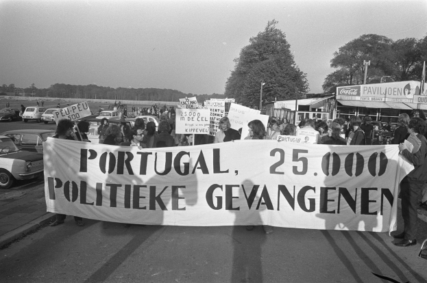 Eine Gruppe von Menschen hält ein Banner mit der Aufschrift "Portugal 25.000 politische Gefangene" auf einer Straße mit parkenden Autos und Bäumen im Hintergrund.
