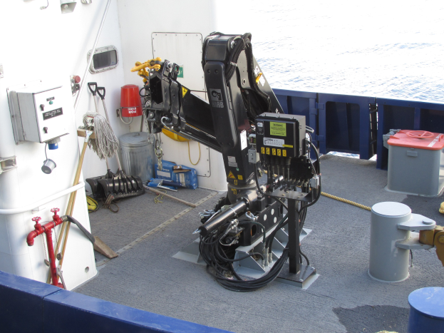 Roboterarm auf einem Bootdeck umgeben von Maschinen und Ausrüstung, mit Wasser im Hintergrund.