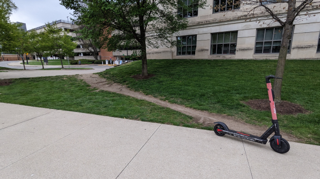 Electric scooter parked on a sidewalk in front of a building, with grass, trees, poles, and other objects nearby, under a visible sky.