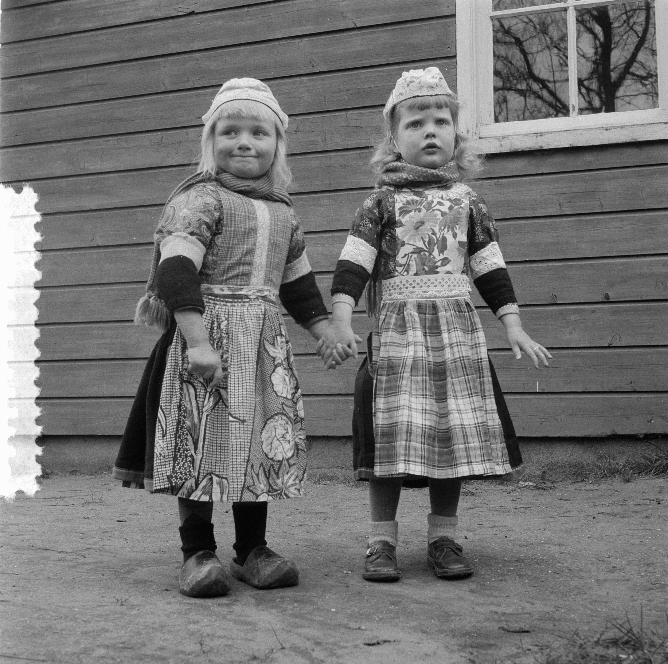 Zwei junge Mädchen in Kleidern und Mützen stehen Hand in Hand vor einem Haus mit einer Wand und einem Fenster, auf einem Schwarz-Weiß-Bild.