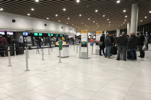 Menschen stehen in einer Schlange am Flughafen, warten in der Nähe von Schildern und Bildschirmen, einige halten Taschen.