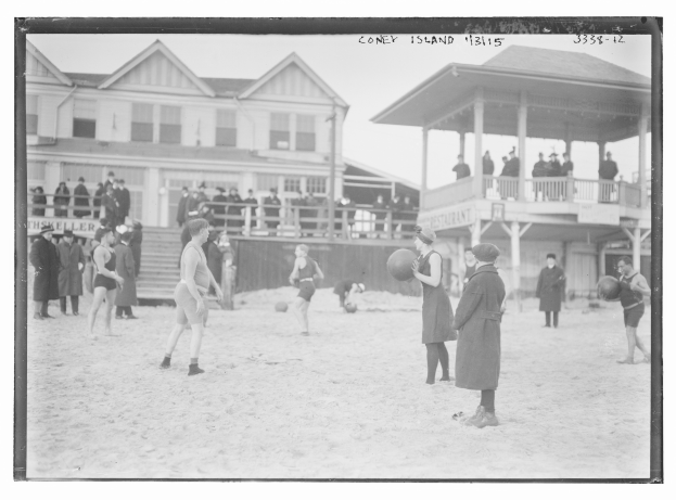 Schwarzes und weißes Foto von Menschen, die Fußball am Strand spielen, mit Gebäuden und Himmel im Hintergrund.