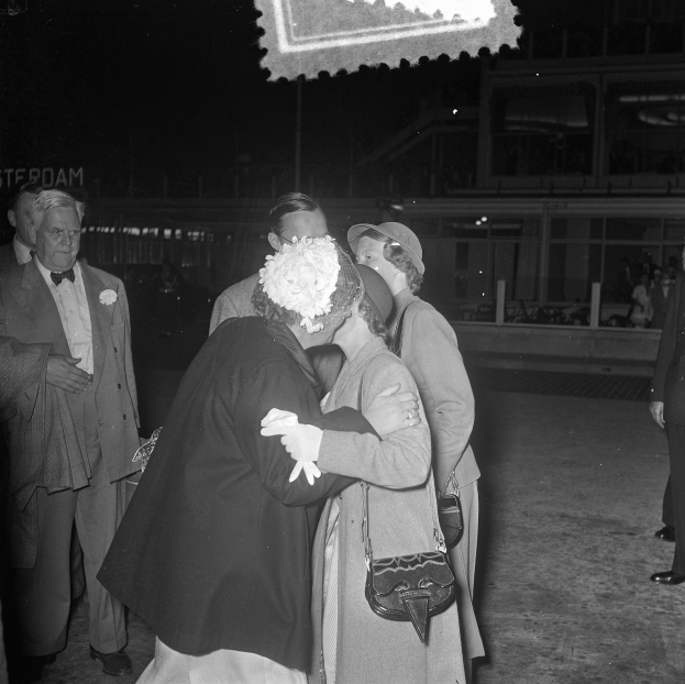 Schwarzes und weißes Foto eines Paares, das sich in einer Menschenmenge umarmt und küsst, umgeben von Menschen und einem Gebäude im Hintergrund.