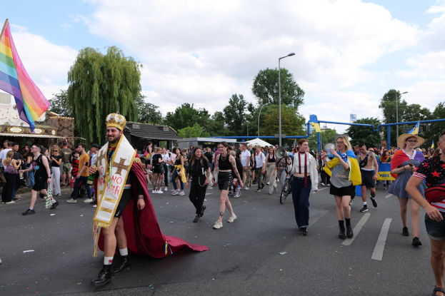 Eine Gruppe von Menschen marschiert bei der Gay Pride Parade 2018 mit einer Regenbogenfahne und Musikinstrumenten, während im Hintergrund Laternenpfähle, Bäume, Schuppen und ein bewölkter Himmel zu sehen sind.