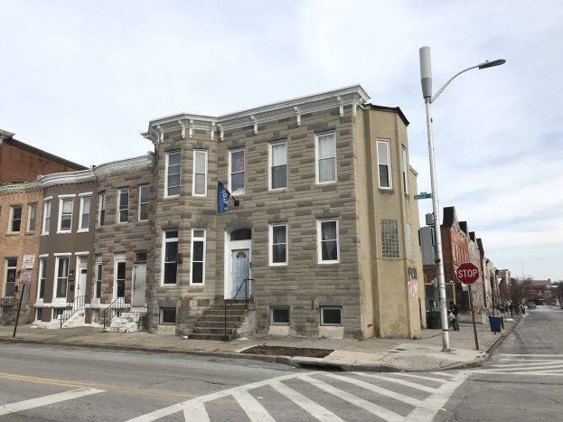 Eine Häuserreihe an einer Straßenecke in einem neuen Wohnprojekt in Philadelphia, mit Fenstern, Türen, Geländern, Laternen, Schildern und Fahrzeugen, sowie Bäumen und einem bewölkten Himmel im Hintergrund.