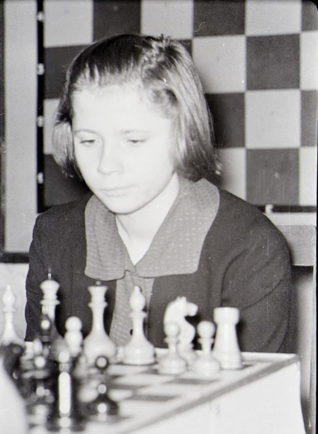 Ein junges Mädchen sitzt an einem Schachbrett, seine Figuren sind in Startpositionen auf einem schwarz-weißen Brett angeordnet.