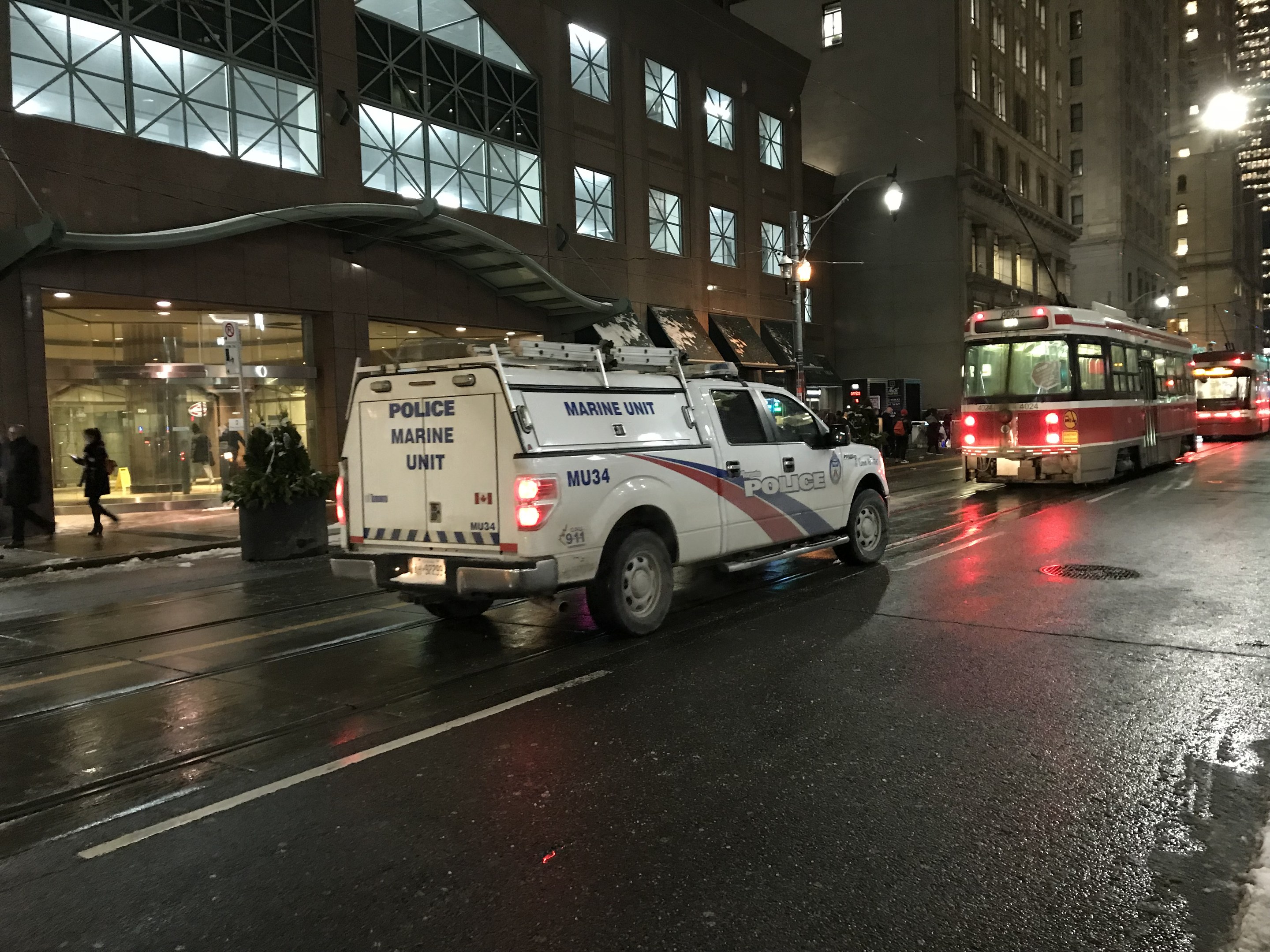 Ein Polizei-Lkw parkt nachts am Straßenrand, mit Fahrzeugen auf der Straße, Fußgängern auf dem Gehweg, Topfpflanzen, Straßenlaternen, Gebäuden mit Fenstern und einem bewölkten Himmel.