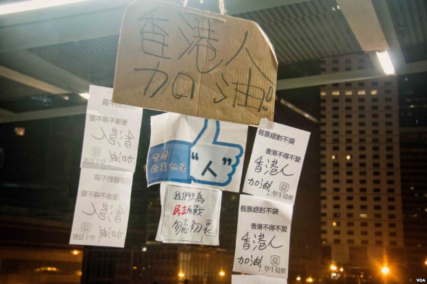 Gruppe von Menschen, die vor einem beleuchteten Gebäude an einem Abend mit Protestschildern an der Decke stehen.