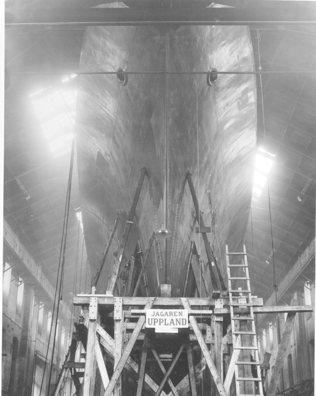 Black and white photo of a large boat under construction in a shipyard, featuring a ladder, wooden poles, a signboard, metal frames, ceiling lights, and windows.