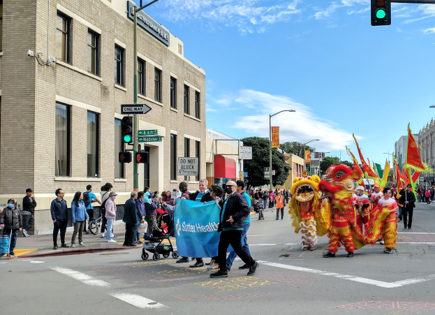 Eine Gruppe von Menschen geht auf einer Straße in San Francisco während des chinesischen Neujahrsfestes, einige in Kostümen mit Fahnen, andere schieben Kinderwagen, mit Gebäuden, Laternenmasten, Verkehrsampeln, Texttafeln, Bäumen und Wolken im Hintergrund.