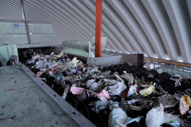 Großer Haufen Müll auf einem Förderband in einer Recyclinganlage, mit Maschinen und einem Metalldach.