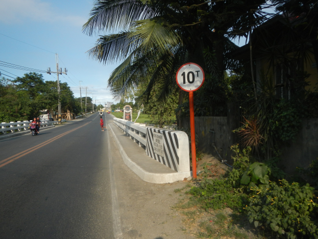 Speed limit sign on a roadside with motorbikes, trees, plants, grass, a fence, electric poles with wires, and a cloudy sky in the background.