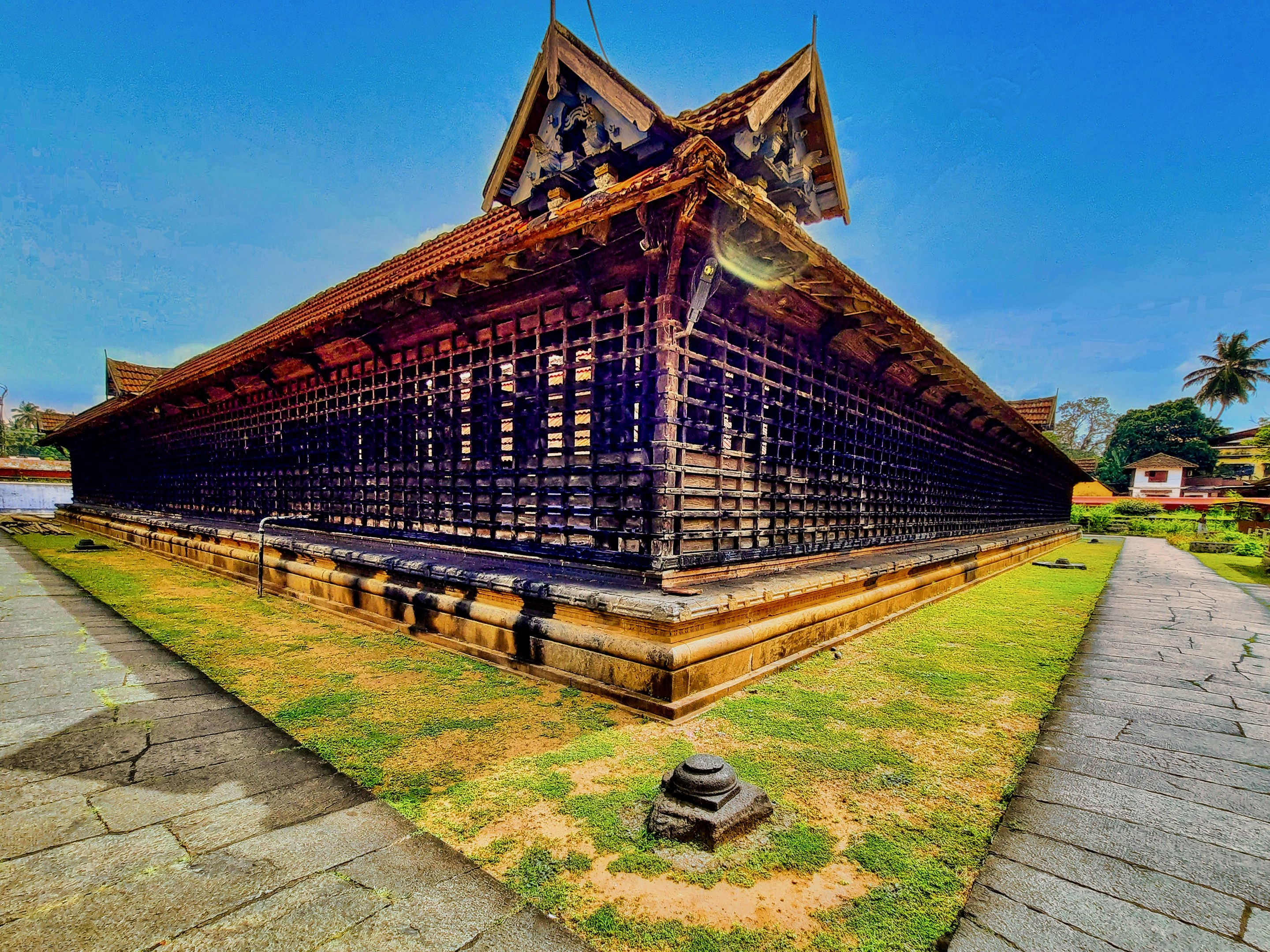 Großes mehrstöckiges Tempeltempelgebäude mit zahlreichen Fenstern, umgeben von Gras und einem gepflasterten Weg, vor Bäumen, Häusern und einem klaren blauen Himmel in Kerala, Indien.