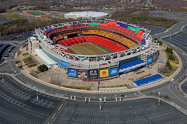 Luftaufnahme eines Footballstadions in einer städtischen Umgebung, umgeben von Gebäuden, Bäumen, Straßen, Fahrzeugen, Masten und Werbetafeln.