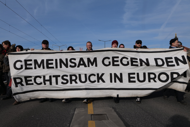 Eine Gruppe von Menschen auf der Straße mit einem Banner, auf dem "Gemeinsam Gegen den Rechtsruck in Europa" steht, mit Straßenlaternen, Strommasten, Stromkabeln, Gebäuden und einem bewölkten Himmel im Hintergrund.
