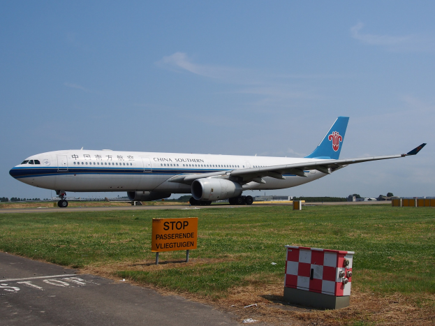 Ein Boeing 787-9 Dreamliner der China Southern Airlines auf dem Rollfeld, umgeben von Gras, einer Texttafel, Gebäuden, Bäumen und einem klaren blauen Himmel.