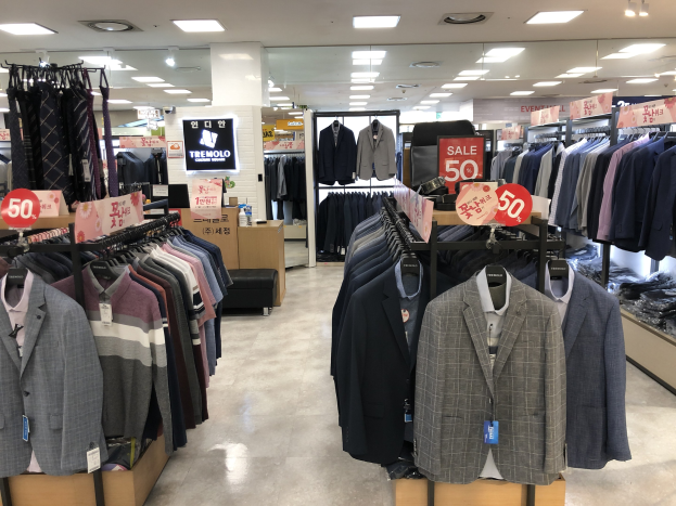 Ein Kleiderladen mit zahlreichen Anzügen an der Stange mit Preisschildern, Deckenleuchten, einer Säule und einem Schild mit der Aufschrift "Tremolo" im Hintergrund.