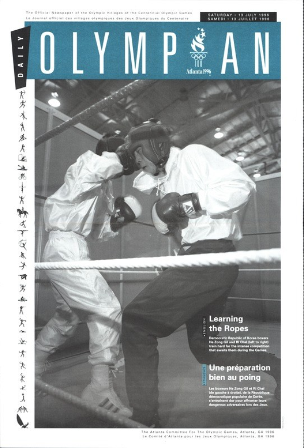 Schwarzes und weißes Foto von zwei Boxern in einem Ring mit Helmen und Handschuhen, umgeben von Seilen und beleuchtet von Überkopfleuchten, mit 'Olympian Magazine'-Text oben und unten.