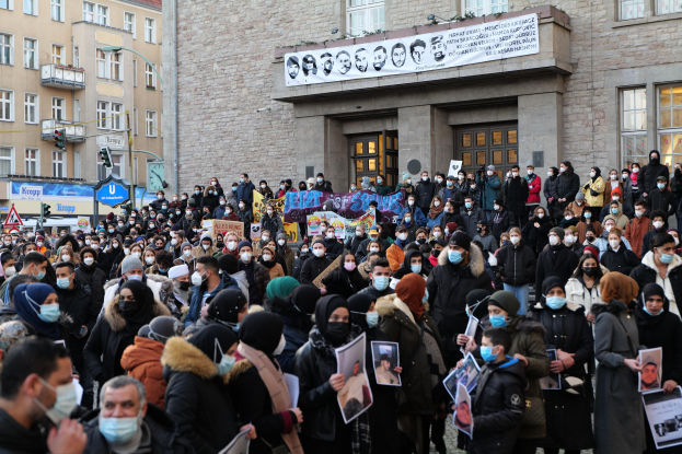 Eine große Gruppe von Menschen mit Masken und Schildern, die vor einem Gebäude mit Bannern, Laternenmasten und Verkehrszeichen protestieren.